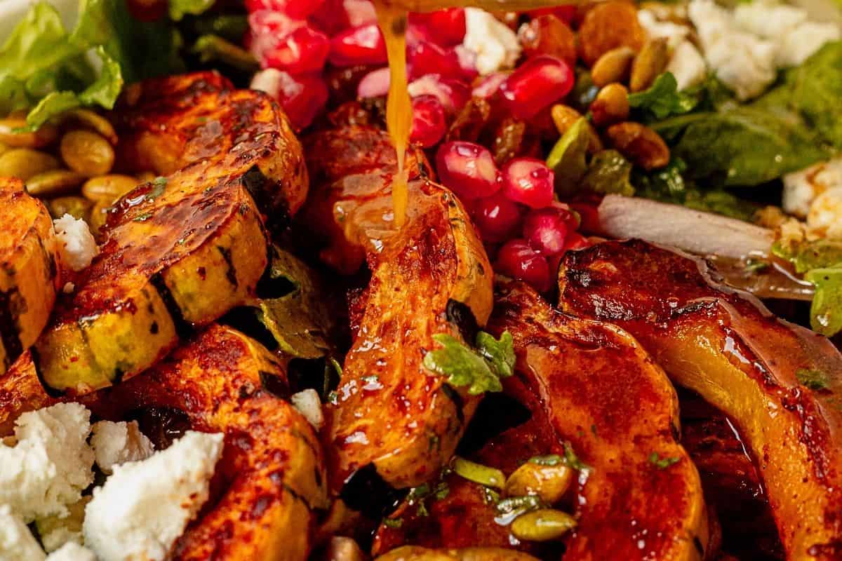 pouring apple cider vinaigrette over roasted delicata saquash, kale, goat cheese, and pomegranates