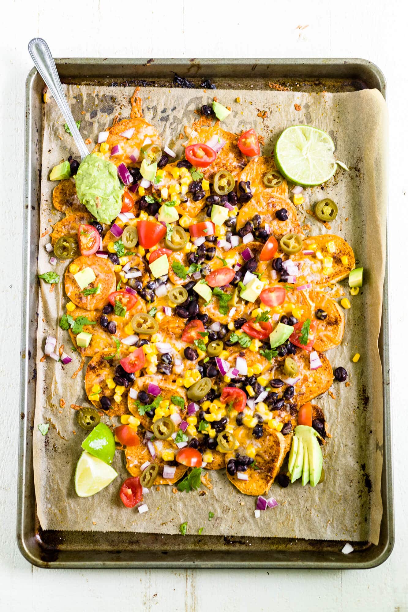 sweet potato nachos on a sheet pan topped with jalapenos, corn, beans, avocado, tomatoes and cilantro