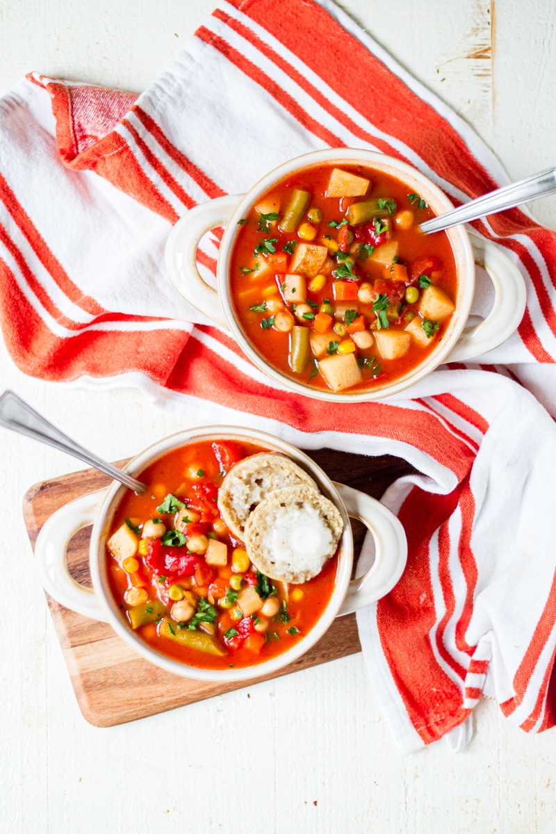 two bowls of homemade vegetable soup on a wood board with a white and orange towel and spoons