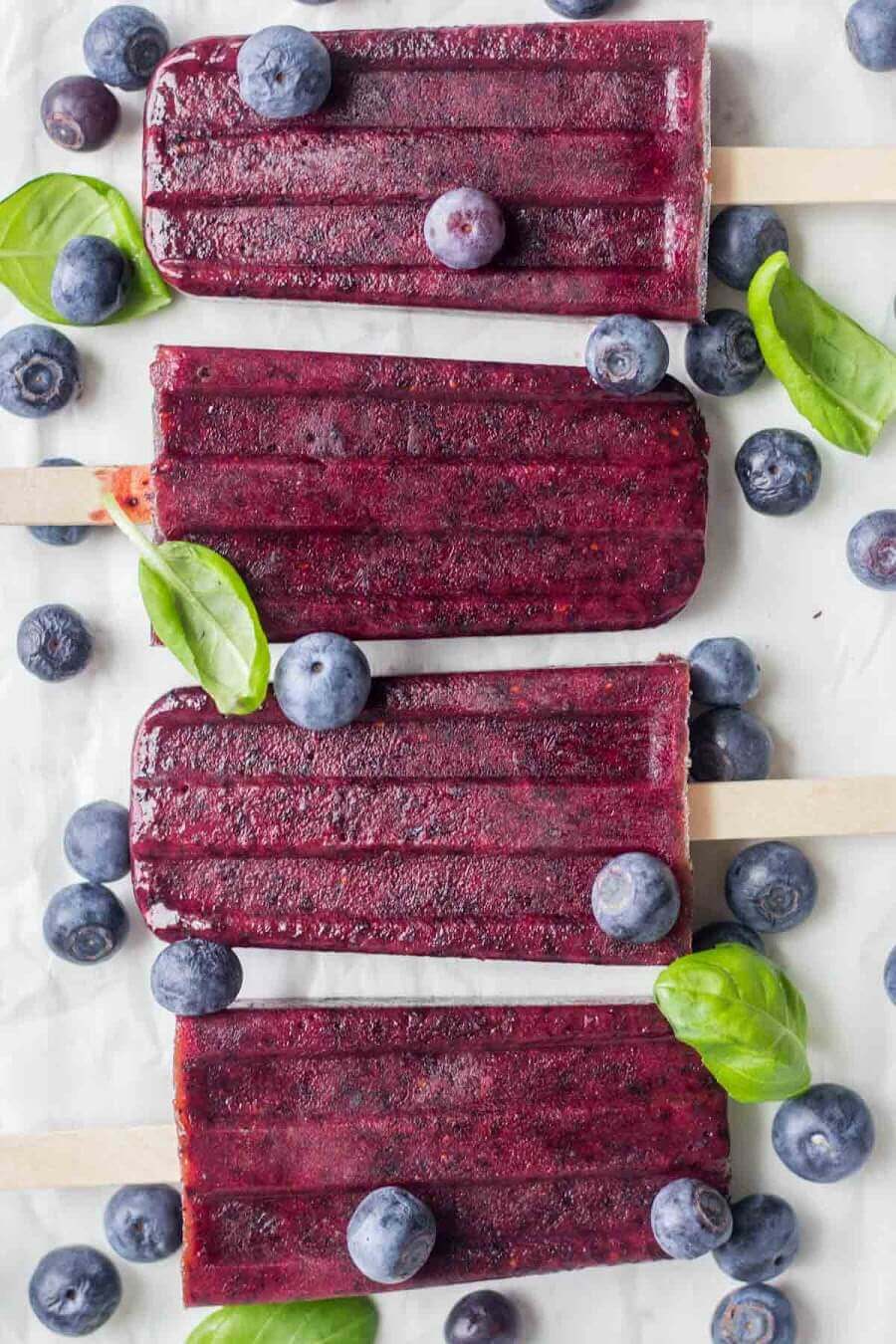 four homemade blueberry popsicles laying on parchment paper surrounded by fresh blueberries and basil