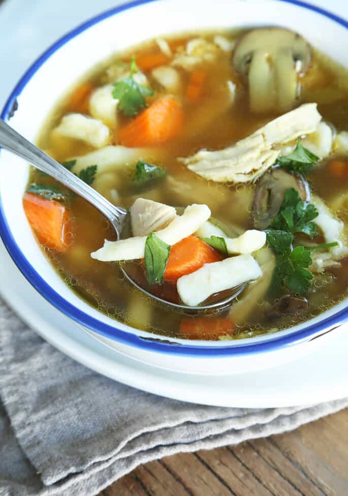 Chicken broth with cooked carrots, mushrooms, chicken, and spaetzle in a white bowl with a metal soup spoon.