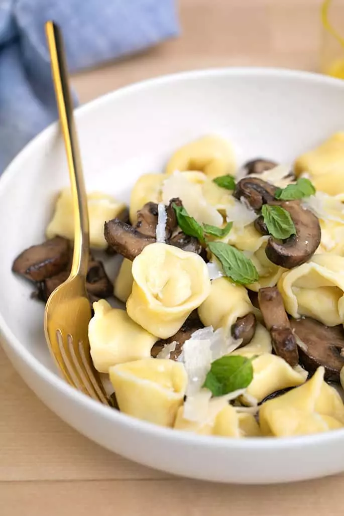 Cooked tortellini with mushrooms, basil, and parmesan shavings with gold fork in white bowl