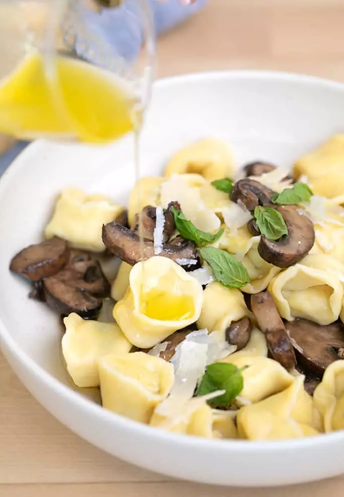 Melted garlic butter pouring onto tortellini with cooked mushrooms in white bowl