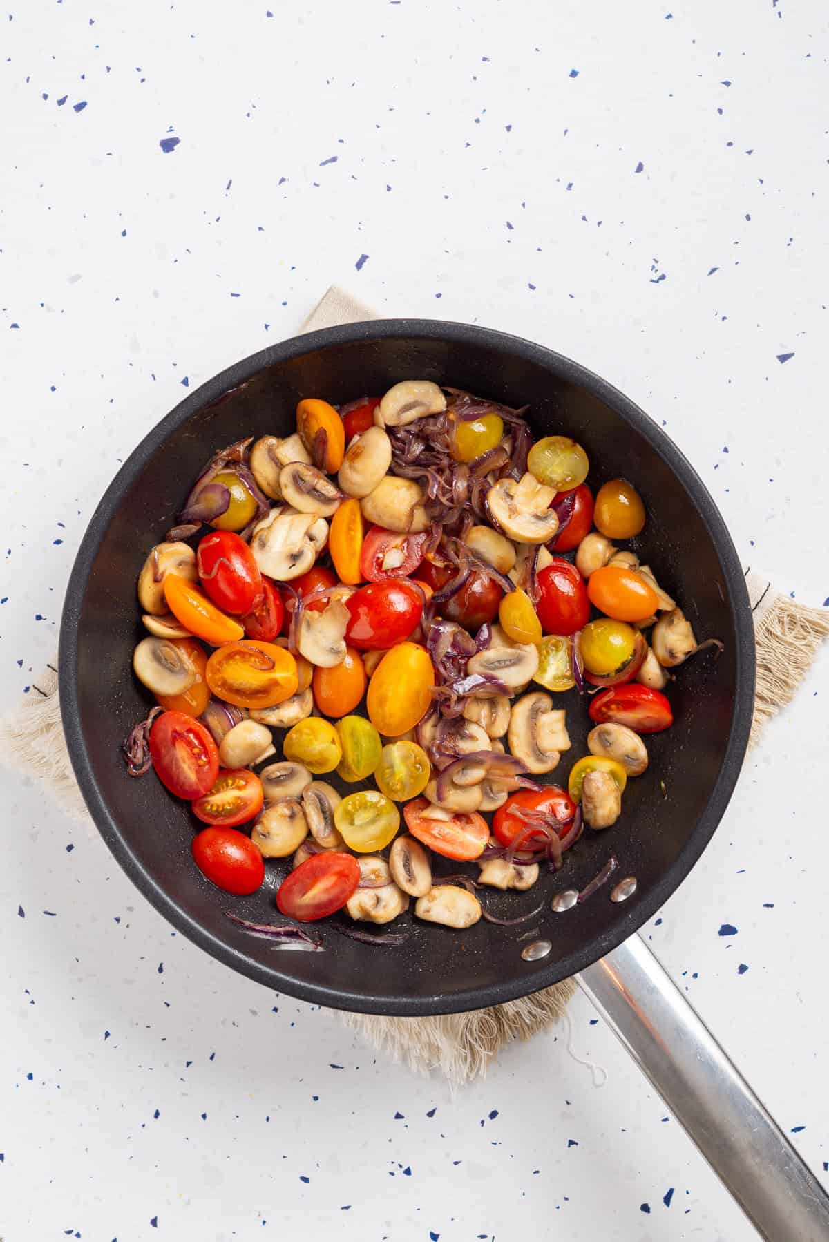 An overhead image of onions, mushrooms, and cherry tomatoes in a skillet.