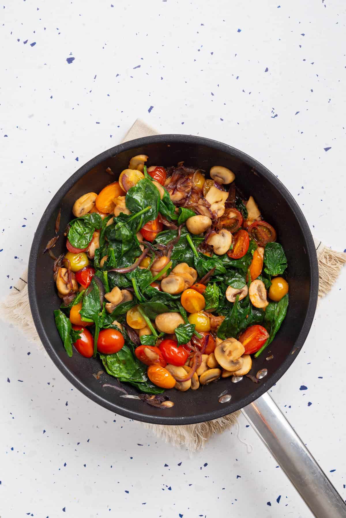 An overhead image of onions, mushrooms, cherry tomatoes, and baby spinach in a skillet.