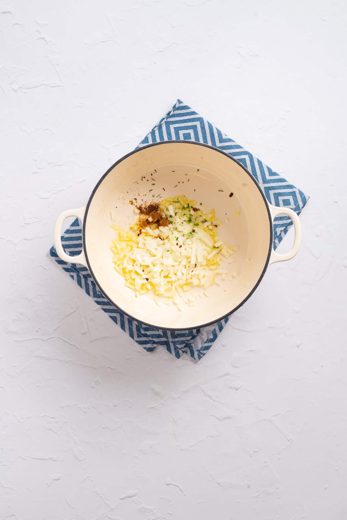 An overhead image of onion, garlic, red pepper flakes, cumin powder, and fresh rosemary being sauted in a dutch oven.